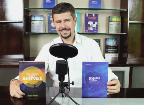 ft sentado sem terno segurando livros sra e en por na frente do microfone e atras estante com livros